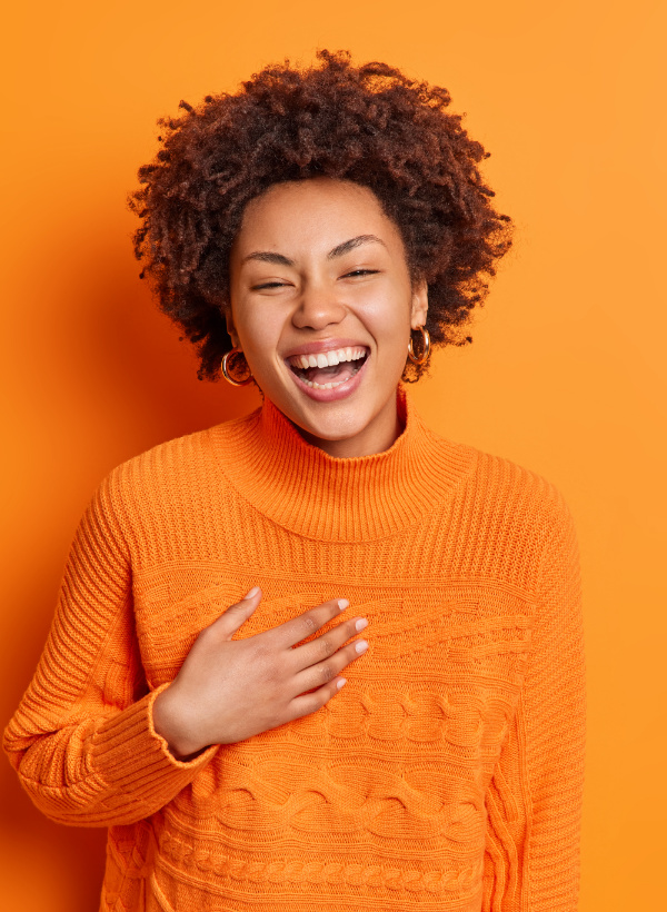 une femme au pull orange et la main sur le coeur rit aux éclats